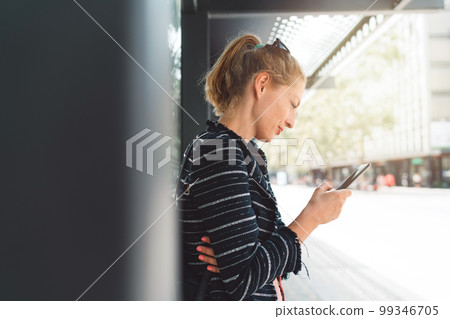 Side profile of caucasian blonde woman staring at her phone while waiting for her bus 99346705