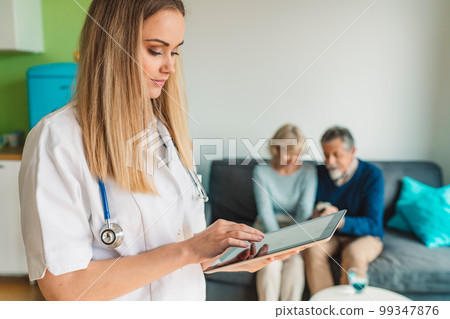 Caucasian nurse looking at the digital tablet, while blurred in the background senior couple sits on the couch and waits for her 99347876