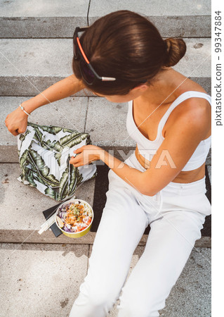 Vertical photo of young woman sitting down on the stairs in the city with her lunch next to her, ready to eat  99347884