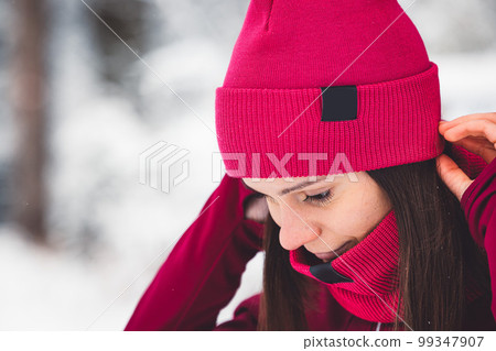 Close up woman putting on a red hat matching her red scarf, while on a walk trough the snowy forest 99347907