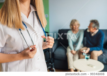 Unrecognizable woman nurse holding for her stethoscope while senior couple is sitting on the couch blurred in the background Unrecognizable woman nurse holding for her stethoscope while senior couple is sitting on the couch blurred in the background 99348008