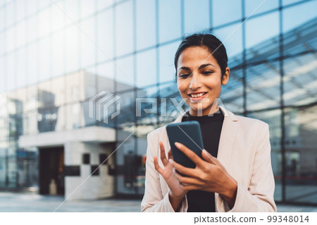 Smiling young business woman writing an email on her phone while standing outside on the sun during her lunch break  99348014