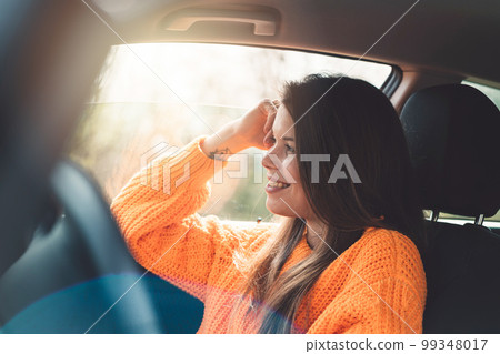 Side profile with sun flare, young smiling woman sitting in her car wearing an orange sweater Side profile with sun flare, young smiling woman sitting in her car wearing an orange sweater 99348017