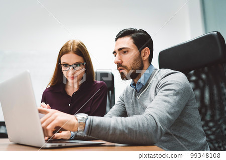 Business man and business woman working on a joint project, sitting by the desk looking at the laptop screen Business man and business woman working on a joint project, sitting by the desk looking at the laptop screen 99348108