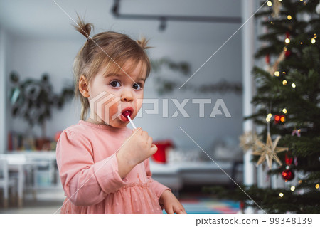 Beautiful caucasian baby gir with two little ponytails holding a Christmas ornament, looking at it with curiosity Beautiful caucasian baby gir with two little ponytails holding a Christmas ornament, looking at it with curiosity 99348139