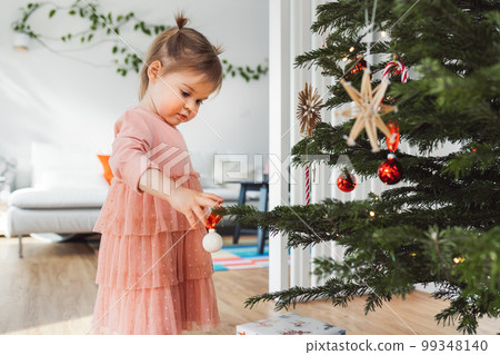 Baby girl wearing a beautiful pink dress putting an ornament on the Christmas tree 99348140
