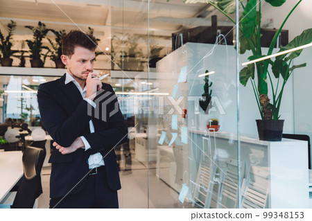 Young business man holding a white pen looking at the clear board where he wrote some ides down  99348153