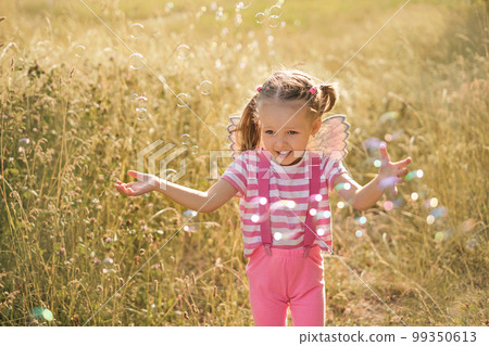 Funny girl with wings on her back runs through the meadow and catches soap bubbles at sunset Funny girl with wings on her back runs through the meadow and catches soap bubbles at sunset 99350613