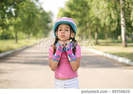 A little beautiful girl fastens a protective helmet. A child rides a skateboard in the park on a summer day 99350620
