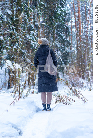 Woman standing her back in winter forest during holiday stroll, walk in woods alone with tote bag, in knitted hat, puffer jacket 99352386