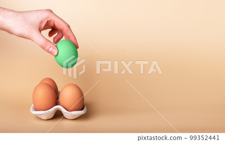 Banner with hand putting outstanding Easter egg in carton box with natural ones on beige background. Green symbol of religious holiday. Standing out from crowd concept. Copy space 99352441