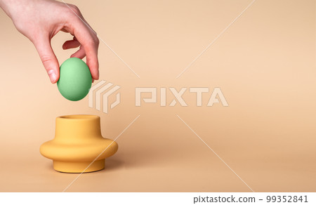 Banner with hand putting green dyed Easter egg in cup. Egg holder and server on beige background. Symbol of Easter holiday. Copyspace 99352841