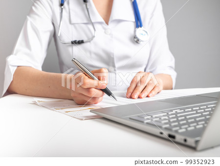 Doctor with stethoscope writing with pen about patient treatment or scheduling appointments. Woman in lab coat sitting at table with laptop. Health care and medicine concept Doctor with stethoscope writing with pen about patient treatment or scheduling appointments. Woman in lab coat sitting at table with laptop. Health care and medicine concept 99352923