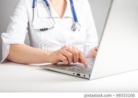 Doctor with stethoscope typing on laptop keyboard. Health care and medicine concept. Woman in lab coat sitting at desk with computer and prescribing medications. 99353100
