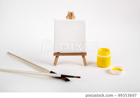 Clean canvas, blank mockup for painting, drawing on wood tripod easel, paint brush and yellow gouache in open jar 99353697