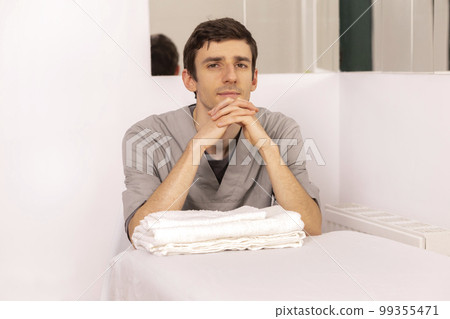 Handsome young male masseur, therapist sits at couch holding hands under chin, looking at camera. Professional therapeutic massage. Mental, health sustainability. Relaxation, healthcare. Horizontal Handsome young male masseur, therapist sits at couch holding hands under chin, looking at camera. Professional therapeutic massage. Mental, health sustainability. Relaxation, healthcare. Horizontal 99355471