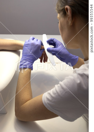 Manicurist polishes cuticle, finger's skin, surface with file. Caucasian manicure master works in gloves at white work station in spa salon. Soft, delicate safe beauty nail care, treatment. Vertical. 99355544