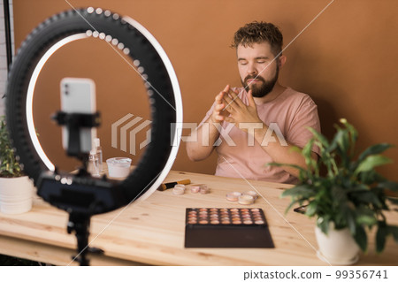 Portrait of happy man beauty blogger sitting in room at table and speaking recommending foundation or decorative cosmetic looking at camera. Blogging and vlog concept 99356741