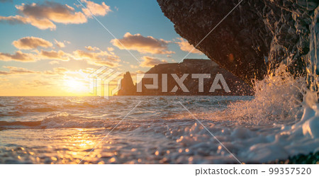 Sea sunset waves with foam. Waves run on pebbles and rock from volcanic basalt. Water breaks into splashes and white foam. Against the background of the cape and the sunset sky. Sea sunset waves with foam. Waves run on pebbles and rock from volcanic basalt. Water breaks into splashes and white foam. Against the background of the cape and the sunset sky. 99357520