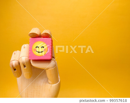 Wooden hand holding pink cube with smile icon on yellow background. Business concept. Wooden hand holding pink cube with smile icon on yellow background. Business concept. 99357618