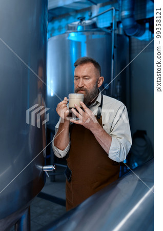 Brewer tasting fresh brewed beer while standing in front of metal containers 99358121