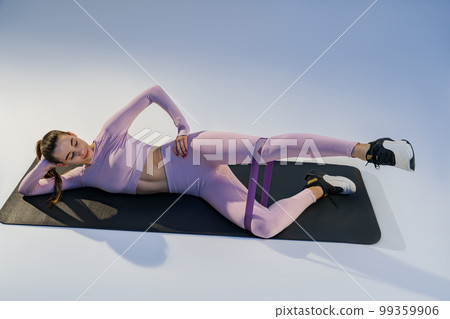 Athletic woman lying on yoga mat and trains legs with fitness elastic band on studio background 99359906