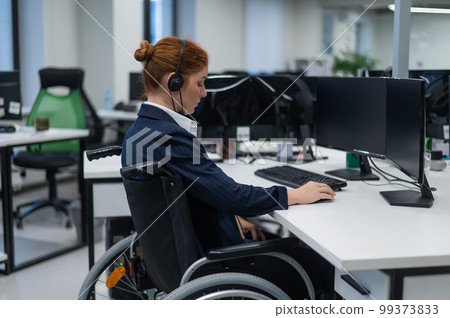 Red-haired caucasian woman in a wheelchair talking on a headset. Female call center worker at her desk. Red-haired caucasian woman in a wheelchair talking on a headset. Female call center worker at her desk. 99373833