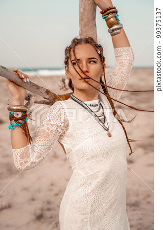 Model in boho style in a white long dress and silver jewelry on the beach. Her hair is braided, and there are many bracelets on her arms. Model in boho style in a white long dress and silver jewelry on the beach. Her hair is braided, and there are many bracelets on her arms. 99375137