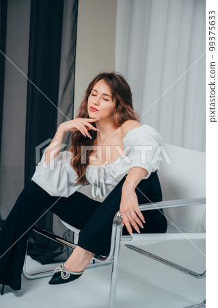 an attractive woman with long hair in a white top and black pants on a chair. 99375633