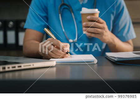 Male medicine doctor, physician or practitioner in lab room writing on blank notebook and work on laptop computer with medical stethoscope on the desk at hospital. Medic tech concept. 99376985