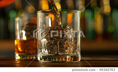 Detail of pouring whiskey into glass with ice cubes. 99380763