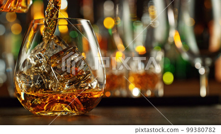 Detail of pouring whiskey into glass with ice cubes. 99380782