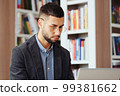Bearded man with eyeglasses wearing jacket browsing internet on laptop in library, doing research and making notes on paper, bookcases on blurred background. Concept of education 99381662