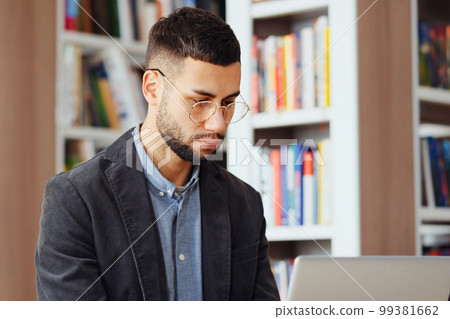 Bearded man with eyeglasses wearing jacket browsing internet on laptop in library, doing research and making notes on paper, bookcases on blurred background. Concept of education Bearded man with eyeglasses wearing jacket browsing internet on laptop in library, doing research and making notes on paper, bookcases on blurred background. Concept of education 99381662