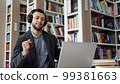 Bearded man with eyeglasses wearing headphones working on laptop in library, starting singing and dancing when hearing popular music. Student taking break in studying and having fun 99381663
