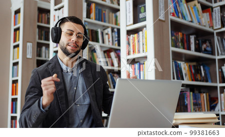Bearded man with eyeglasses wearing headphones working on laptop in library, starting singing and dancing when hearing popular music. Student taking break in studying and having fun 99381663