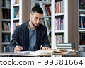 Student wearing eyeglasses writing homework in library, speaking aloud his ideas, heap of books lying beside, bookcases on blurred background. Concept of study 99381664