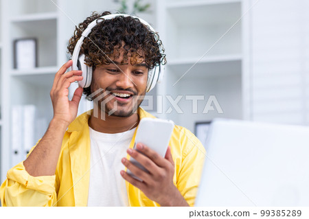 Happy smiling indian man watching video on smartphone close up in yellow shirt in home office, man smiling and surfing online stream using headphones. 99385289