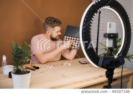 Portrait of happy man beauty blogger sitting in room at table and speaking recommending foundation or decorative cosmetic looking at camera. Blogging and vlog concept Portrait of happy man beauty blogger sitting in room at table and speaking recommending foundation or decorative cosmetic looking at camera. Blogging and vlog concept 99387196