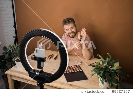 Famous blogger. Cheerful male vlogger showing cosmetics products while recording video and giving advices for his beauty blog. Make-up artist and recording beauty vlog concept Famous blogger. Cheerful male vlogger showing cosmetics products while recording video and giving advices for his beauty blog. Make-up artist and recording beauty vlog concept 99387197