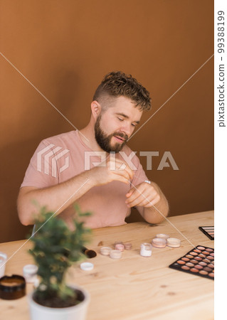 Portrait of happy man beauty blogger sitting in room at table and speaking recommending foundation or decorative cosmetic looking at camera. Blogging and vlog concept 99388199