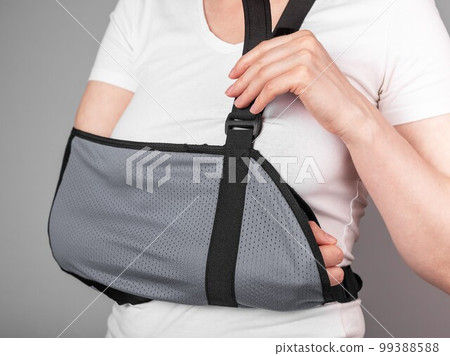 Woman putting on sling to support injured arm. Elbow, shoulder, forearm, wrist pain caused by trauma, injury, chronical disease. Using bandage for ache reduction and immobilizing 99388588