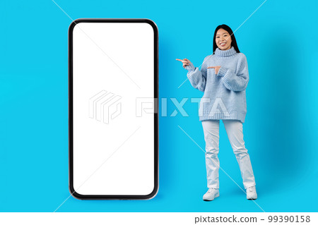 Cheerful korean woman in warm sweater pointing at smartphone, mockup Cheerful korean woman in warm sweater pointing at smartphone, mockup 99390158