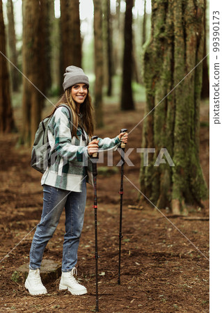 Glad millennial caucasian lady in hat with backpack, trekking sticks, resting in cold forest, enjoy adventure 99390173