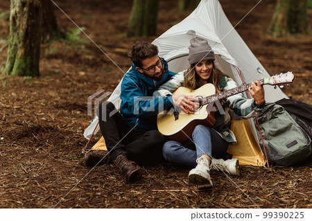 Cheerful millennial caucasian husband and wife in jackets sit in cold forest, enjoy adventure, do lesson, play guitar Cheerful millennial caucasian husband and wife in jackets sit in cold forest, enjoy adventure, do lesson, play guitar 99390225