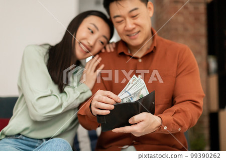 Positive rich middle aged asian man putting banknotes into wallet, sitting with his young wife on sofa at home 99390262