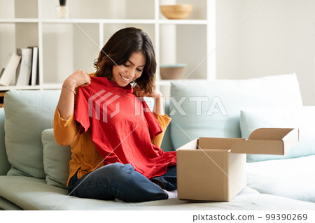 Smiling Young Middle Eastern Female Unpaking Box With New Clothes At Home 99390269