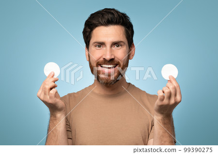 Portrait of excited bearded man with perfect skin holding in hands cotton pads and smiling at camera, blue background 99390275
