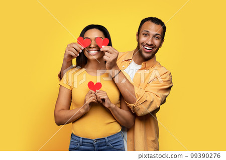 Cheerful young african american man puts hearts to eyes of woman, have fun isolated on yellow background Cheerful young african american man puts hearts to eyes of woman, have fun isolated on yellow background 99390276