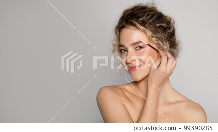 Makeup concept. Attractive woman brushing brows with eyebrow brush, posing over grey background, panorama, free space Makeup concept. Attractive woman brushing brows with eyebrow brush, posing over grey background, panorama, free space 99390285